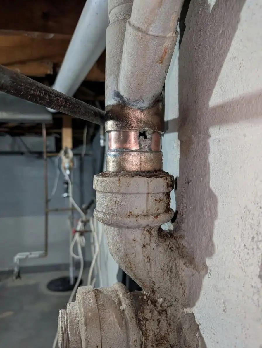 Old cast iron drain stack in an unfinished basement with rust staining and a copper repair section